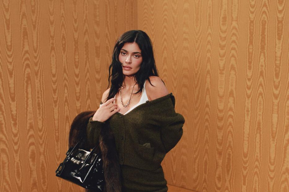 Kylie Jenner posiert für Miu Miu in einem grünen Off-Shoulder-Cardigan und einem passenden Rock dazu. Außerdem trägt sie Kniestrümpfe und schwarze Schuhe zum Schnürren. Ihre Accessoires sind eine goldene Kette, eine kleine Ledertasche und ein brauner Pelzmantel
