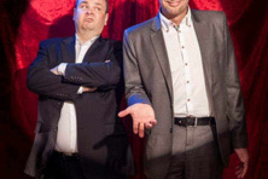 Zwei Männer in Anzügen stehen vor einem roten Vorhang.