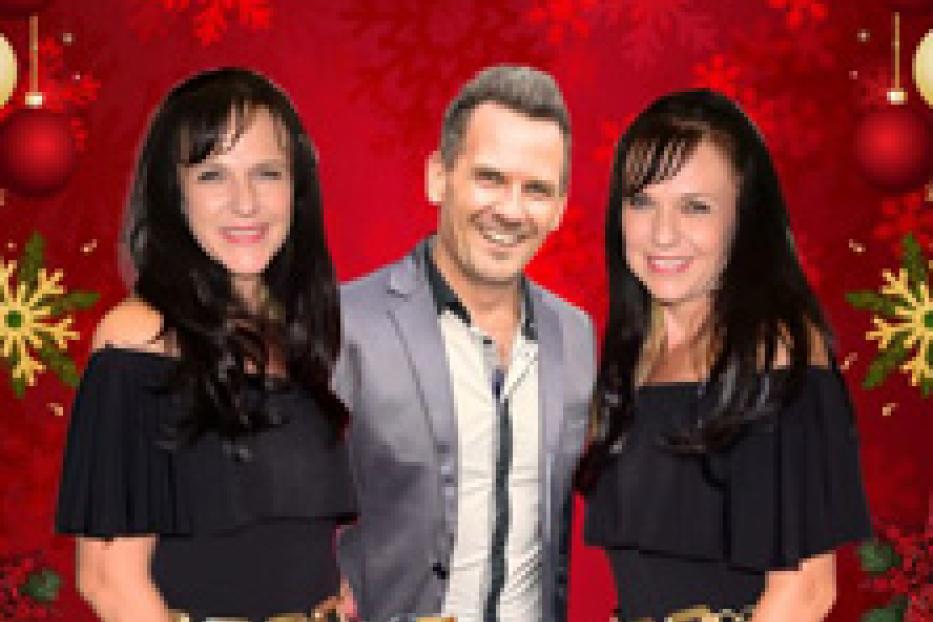 Ein Mann steht zwischen zwei Frauen vor einem weihnachtlichen Hintergrund.