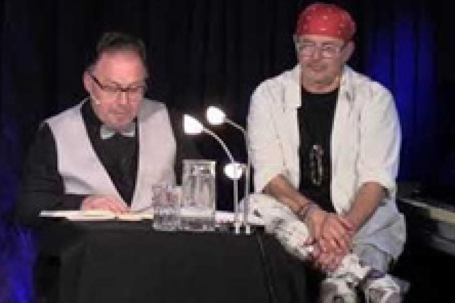 Zwei Männer sitzen an einem Tisch vor einem schwarzen Vorhang.