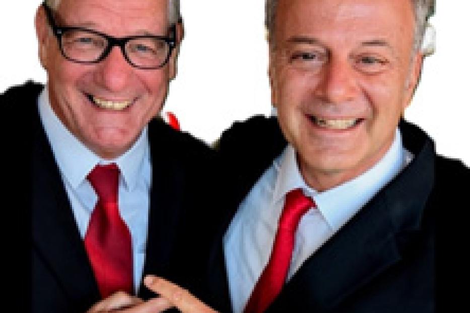 Zwei Männer in Anzügen mit roten Krawatten lächeln in die Kamera.