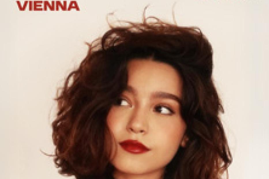 Porträt einer jungen Frau mit dunklen Haaren und rotem Lippenstift vor einem Ankündigungsplakat für Mari Froes in Wien.