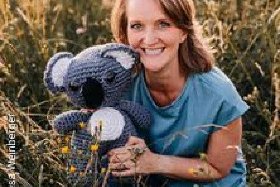 Eine Frau hockt in einem Feld und hält einen gehäkelten Koala-Bären.