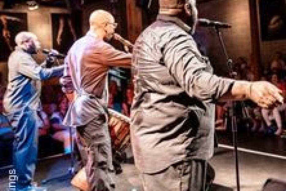 Drei Musiker stehen auf einer Bühne und spielen vor einem Publikum.