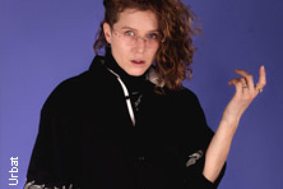 Eine Frau mit lockigem Haar und schwarzer Jacke posiert vor einem blauen Hintergrund.