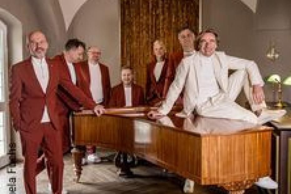 Eine Gruppe von sieben Männern posiert um ein Klavier in einem eleganten Raum.