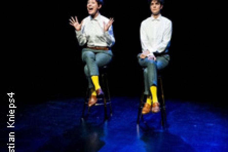 Zwei Personen sitzen auf Stühlen und scheinen auf einer Bühne zu performen.