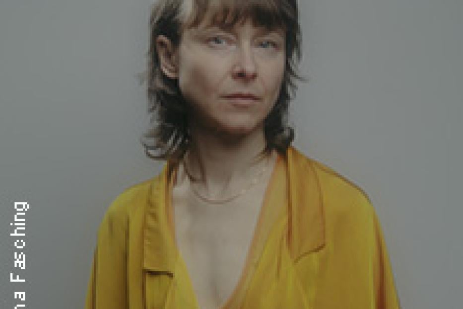 Porträt einer Frau mit blonden Haaren und gelber Bluse vor einem grauen Hintergrund.