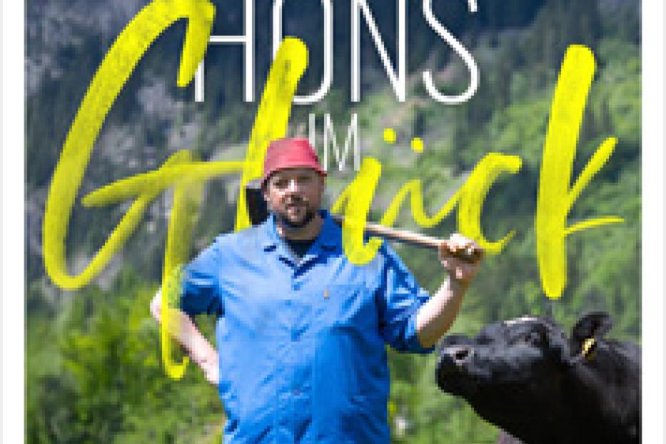 Ein Mann mit Axt und roter Mütze steht vor einer Kuh, im Hintergrund Berge.
