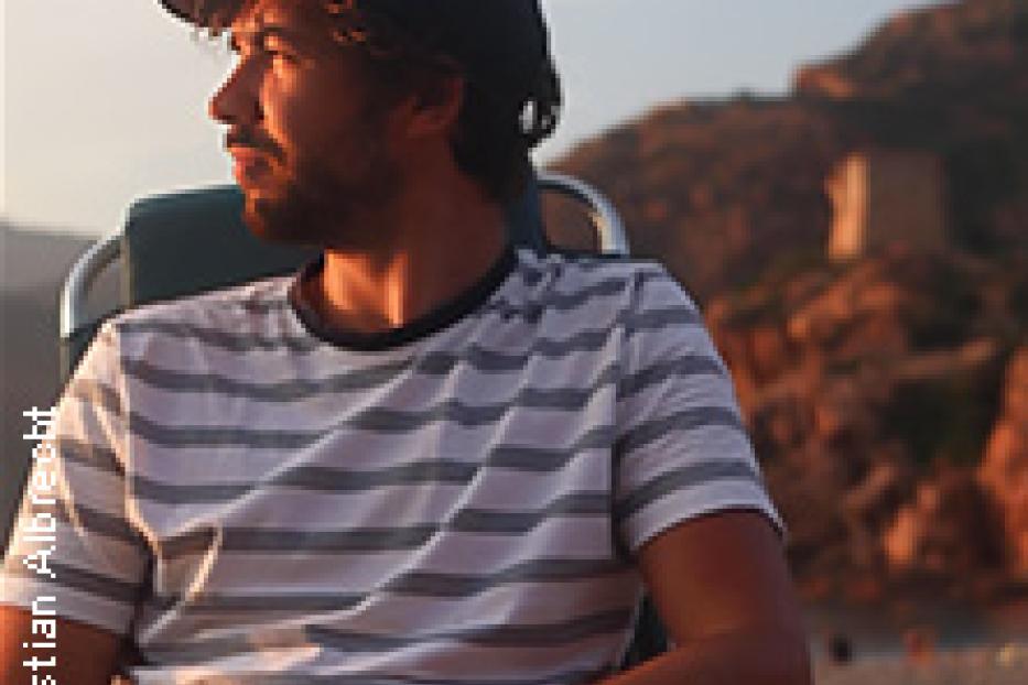 Ein Mann mit Bart und Kappe sitzt am Strand und schaut in die Ferne.