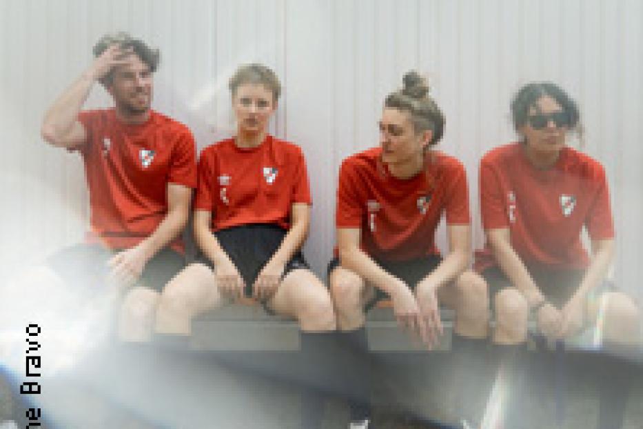 Vier Personen in roten Fußballtrikots sitzen auf einer Bank vor einer weißen Wand.