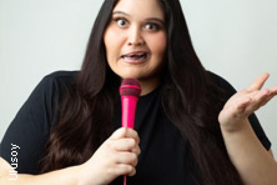 Eine Frau mit langen, dunklen Haaren hält ein pinkfarbenes Mikrofon und spricht.