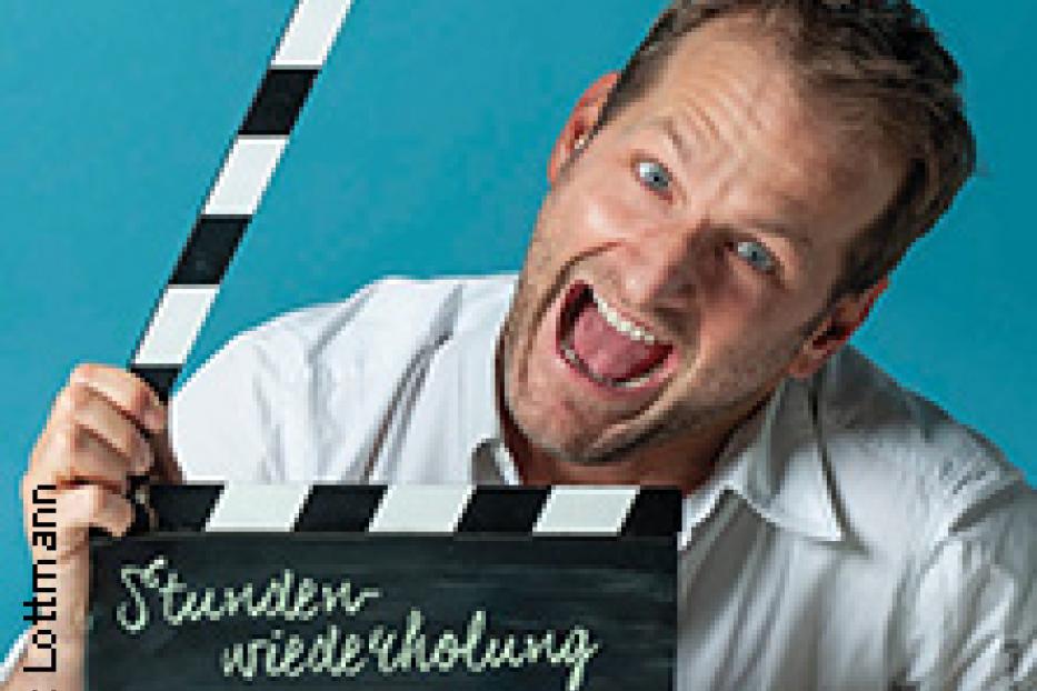 Ein Mann hält eine Filmklappe mit dem Titel „Stundenwiederholung“ in die Kamera.
