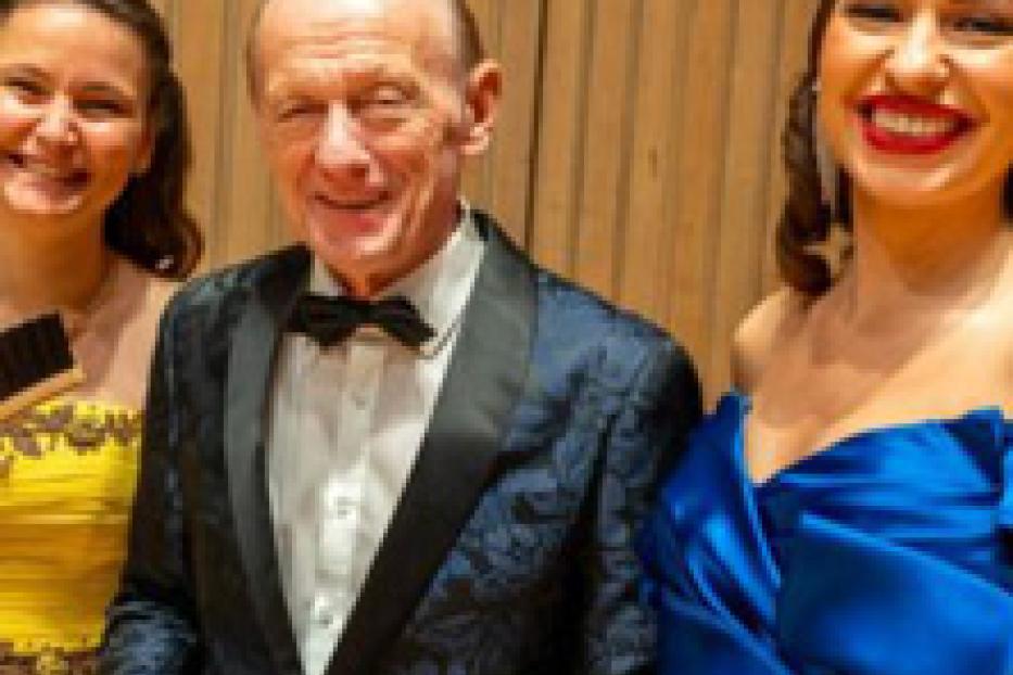 Ein älterer Mann in einem Anzug posiert mit zwei jungen Frauen in Abendkleidern.
