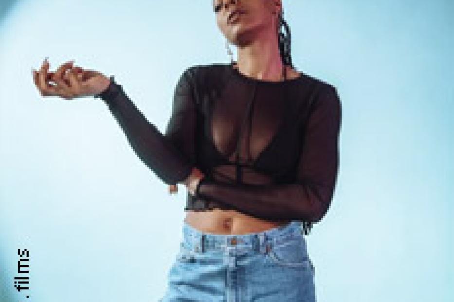 Eine junge Frau mit dunkler Haut und geflochtenen Zöpfen posiert vor einem hellblauen Hintergrund.