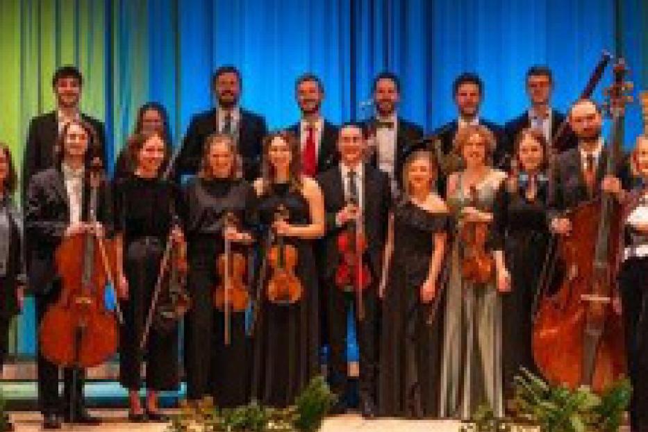Ein Orchester steht auf einer Bühne vor einem blauen Vorhang.