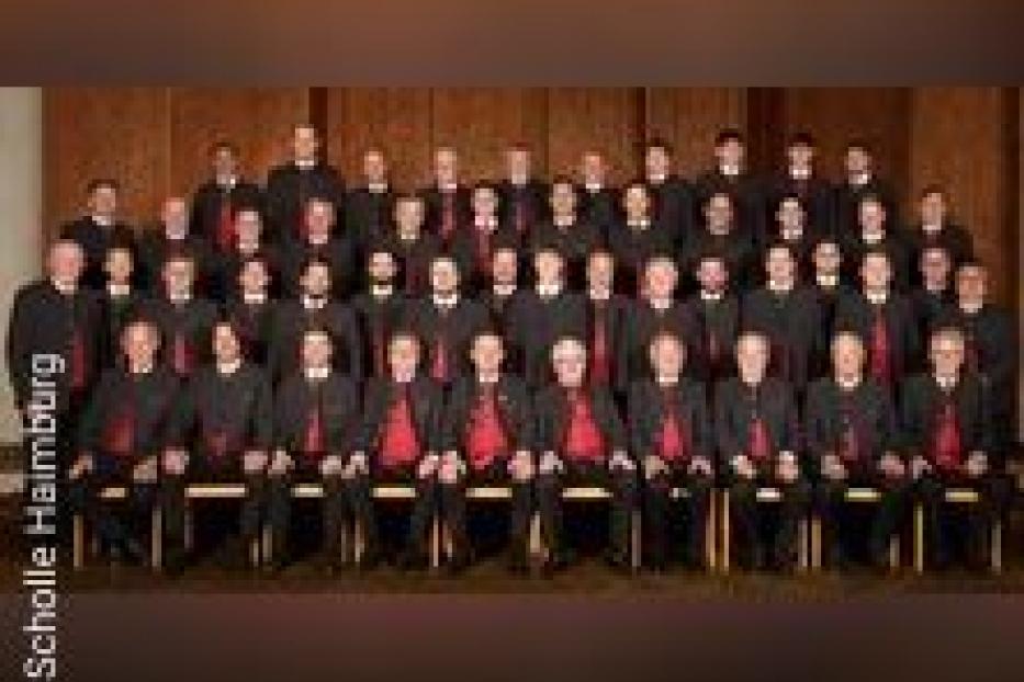 Ein großer Männerchor in schwarzer Kleidung mit roten Akzenten sitzt für ein Gruppenfoto.