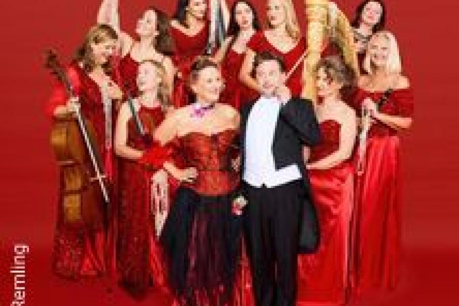 Ein Ensemble aus Frauen in roten Kleidern und ein Mann im Smoking posieren vor einem roten Hintergrund.