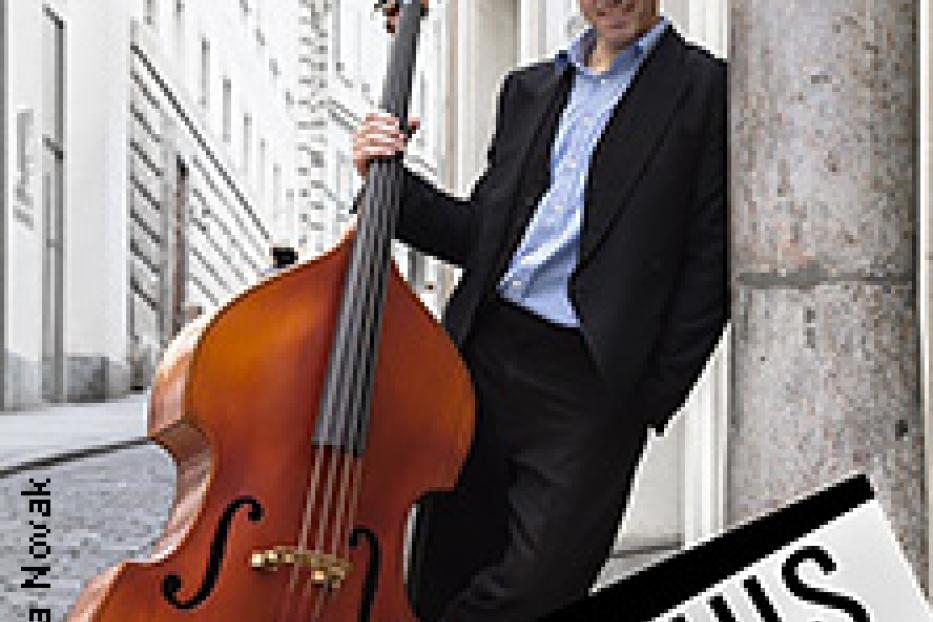 Ein Mann mit Kontrabass lehnt an einer Säule in einer Stadt.