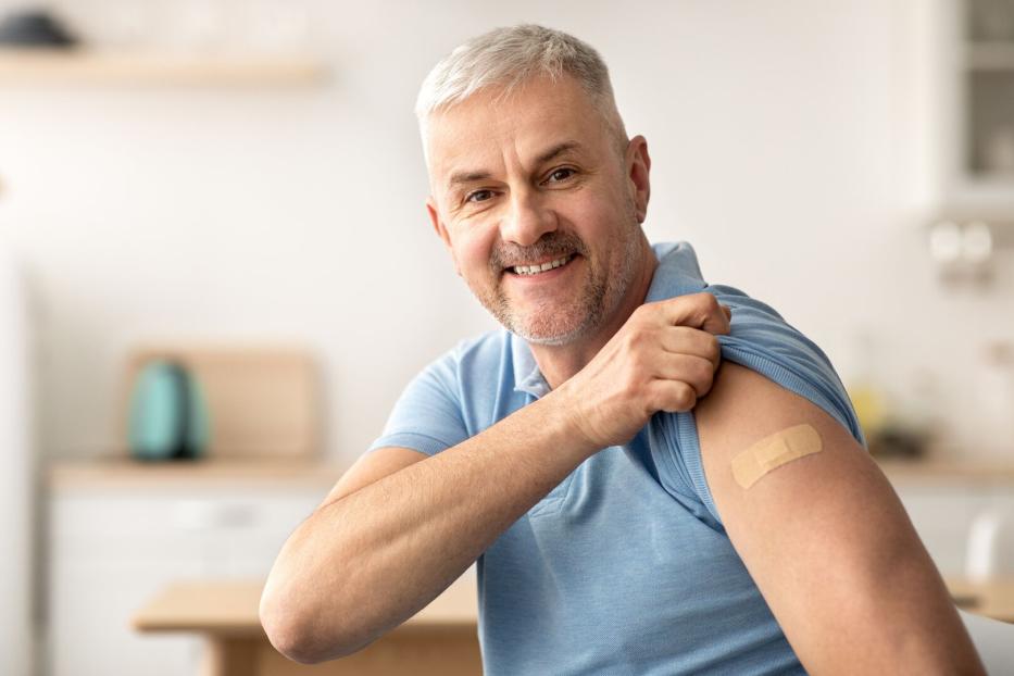 Mann in blauem Poloshirt zeigt Pflaster auf Oberarm, sitzt in heller Küche mit unscharfem Hintergrund.