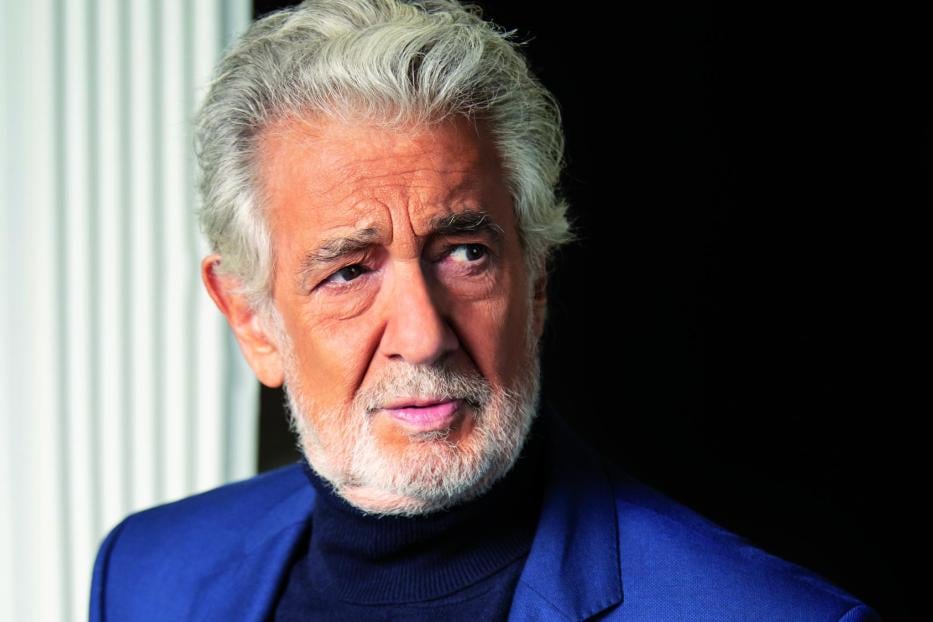 Älterer Mann mit grauen Haaren und Bart trägt einen dunkelblauen Anzug und schwarzen Rollkragenpullover vor schwarzem Hintergrund.