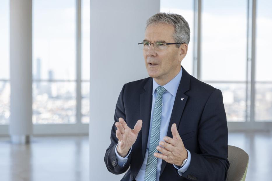 Mann in dunklem Anzug, hellblauem Hemd und gemusterter Krawatte sitzt vor großen Fenstern mit Stadtblick und gestikuliert mit den Händen.