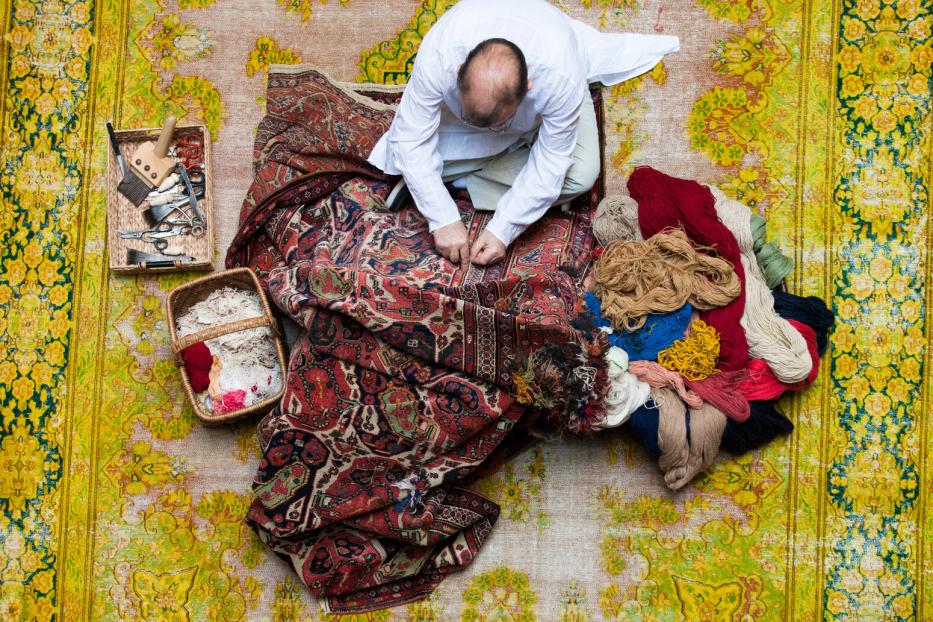 Vogelperspektive: Ein Mann hockt am Boden, vor ihm ein Teppich, neben ihm Wolle und Flickzeug. Er arbeitet am Perser mit klassischem Muster.