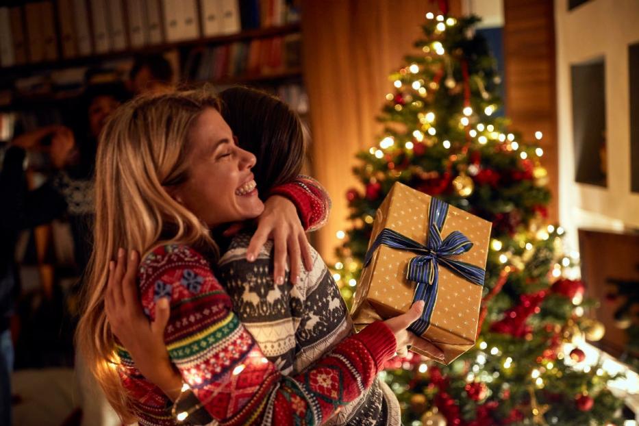 Zwei Frauen umarmen sich und übergeben sich Geschenke vor dem Christbaum in einem weihnachtlich geschmückten Raum.