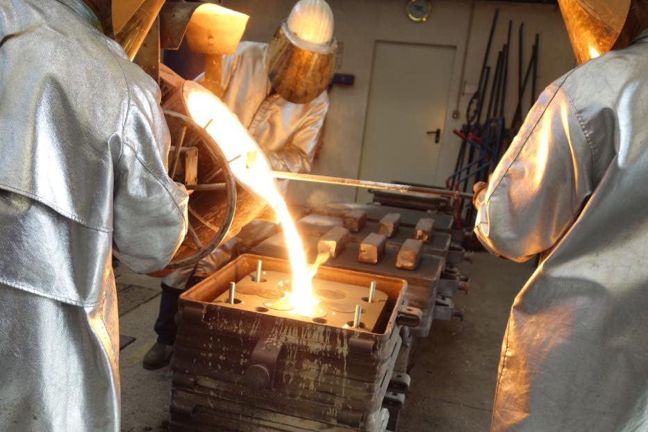 drei Menschen in silbernen Schutzanzügen gießen flüssiges Metall in eine Form