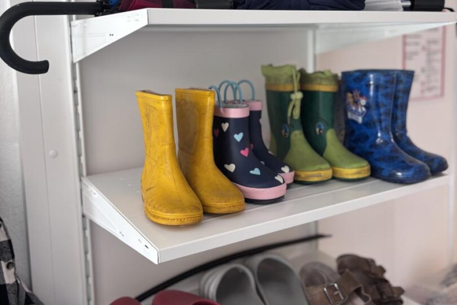 Mehrere Paar Gummistiefel und Sandalen stehen ordentlich auf Regalbrettern, darüber liegen zusammengefaltete Regenschirme.