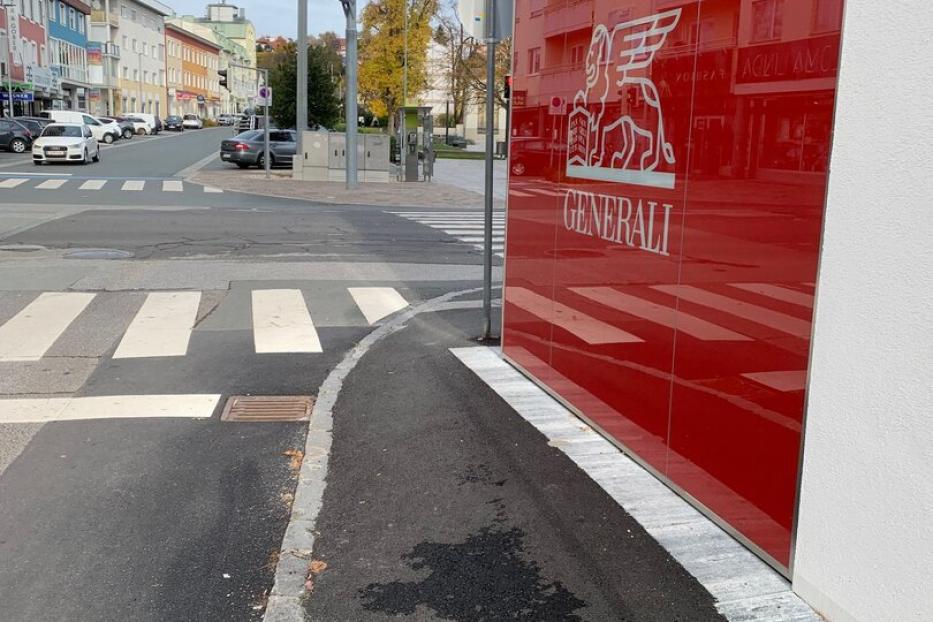 Ein schmaler Gehweg führt an einer roten Wand mit Generali-Logo entlang, daneben eine Kreuzung mit Ampeln und Zebrastreifen.