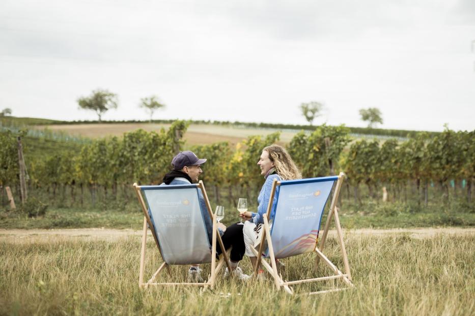 Zwei Menschen sitzen in Liegestühlen auf einer Wiese vor einem Weinberg und unterhalten sich bei einem Glas Wein.