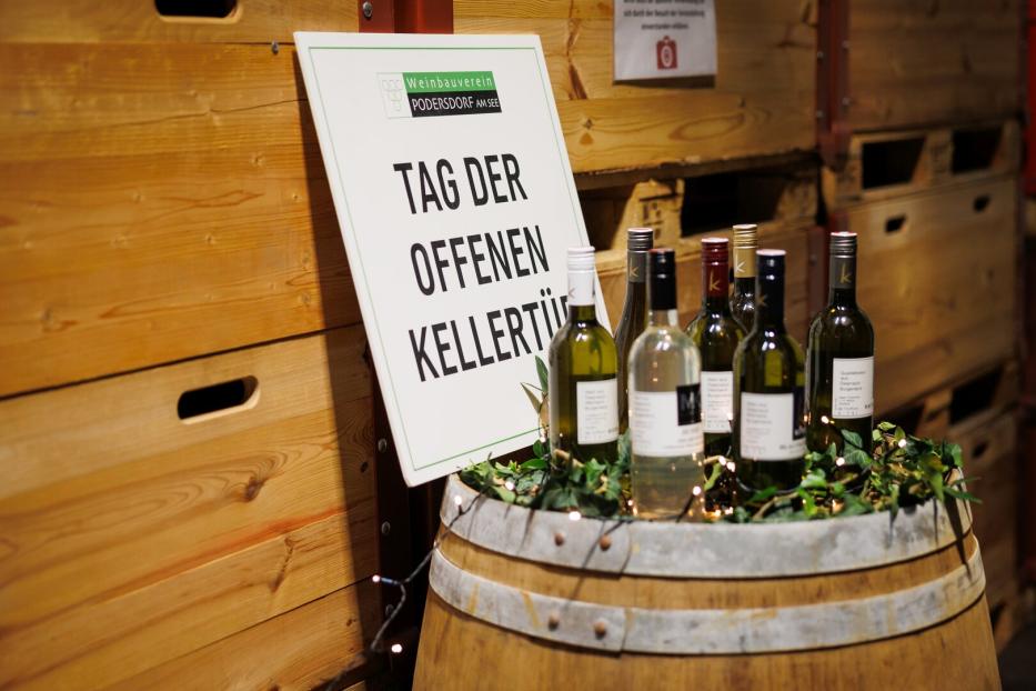 Mehrere Weinflaschen stehen auf einem Weinfass, daneben ein Schild mit der Aufschrift „Tag der offenen Kellertür“.