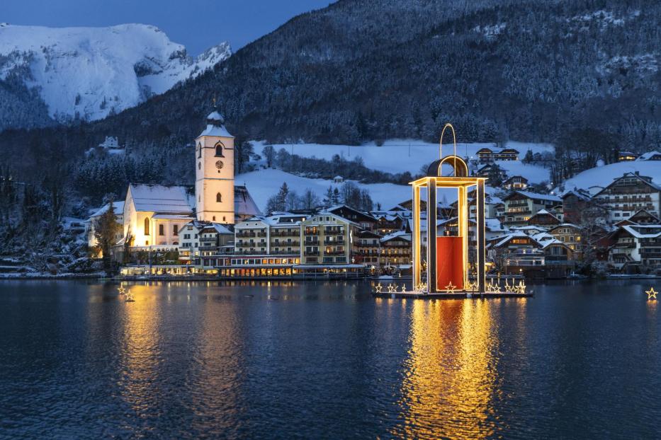 Beleuchtete Kirche und Häuser am verschneiten Seeufer des Wolfgangsees, große schwimmende Weihnachtslaterne auf dem Wasser, Berge im Hintergrund.