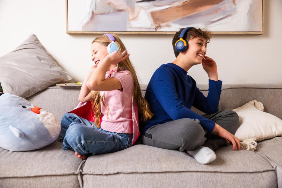 Zwei Kinder sitzen lachend auf einem Sofa und tragen die myFirst CareBuds Max Kopfhörer in verschiedenen Farben.