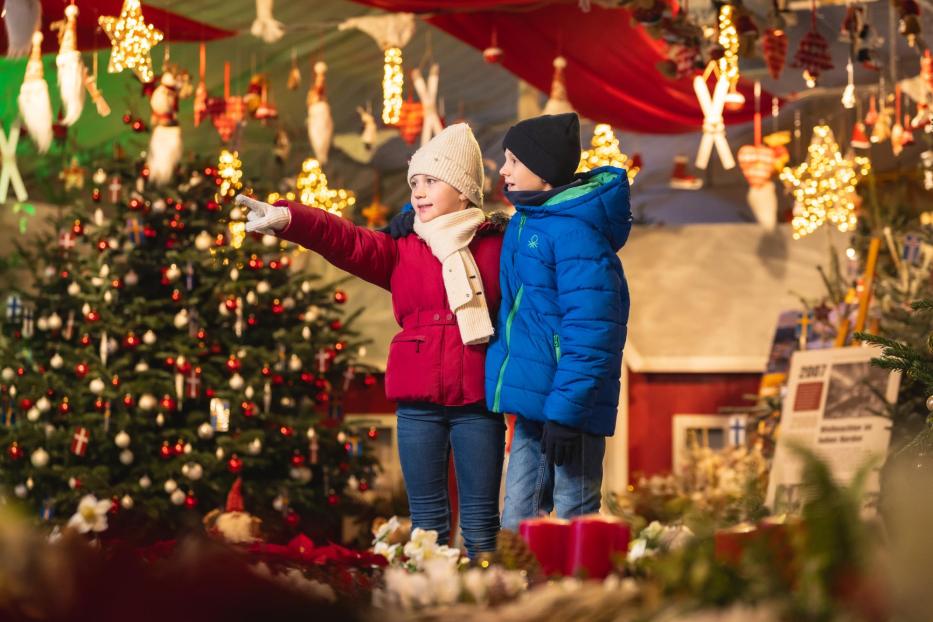 Zwei Kinder in Winterkleidung stehen vor einem Weihnachtsbaum in Hirschstetten und blicken auf einen Weihnachtsmarkt.