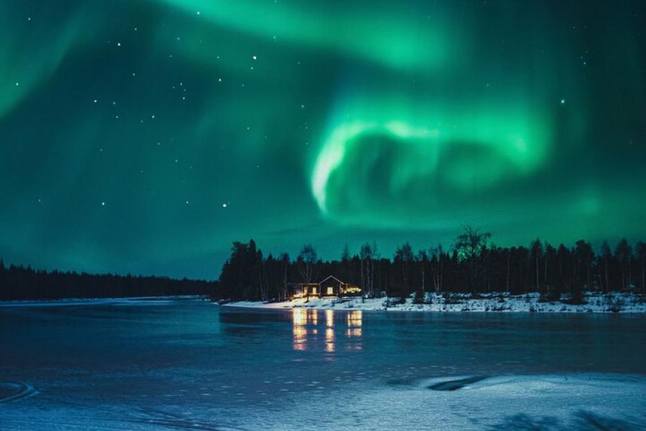 Grünes Polarlicht leuchtet am Nachthimmel über einem zugefrorenen See mit beleuchtetem Haus am Ufer.