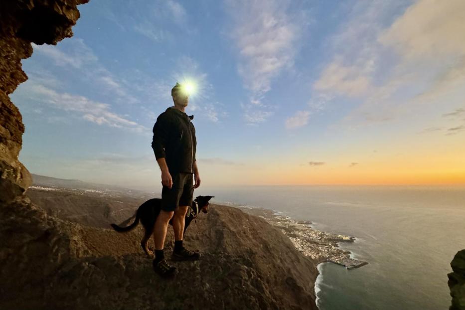Mann mit Stirnlampe und Hund steht auf einer Klippe in der Dämmerung