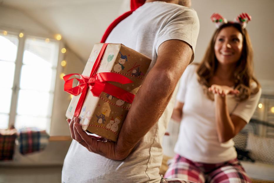 Ein Mann versteckt ein Geschenk mit roter Schleife hinter dem Rücken, während eine Frau erwartungsvoll lächelt.