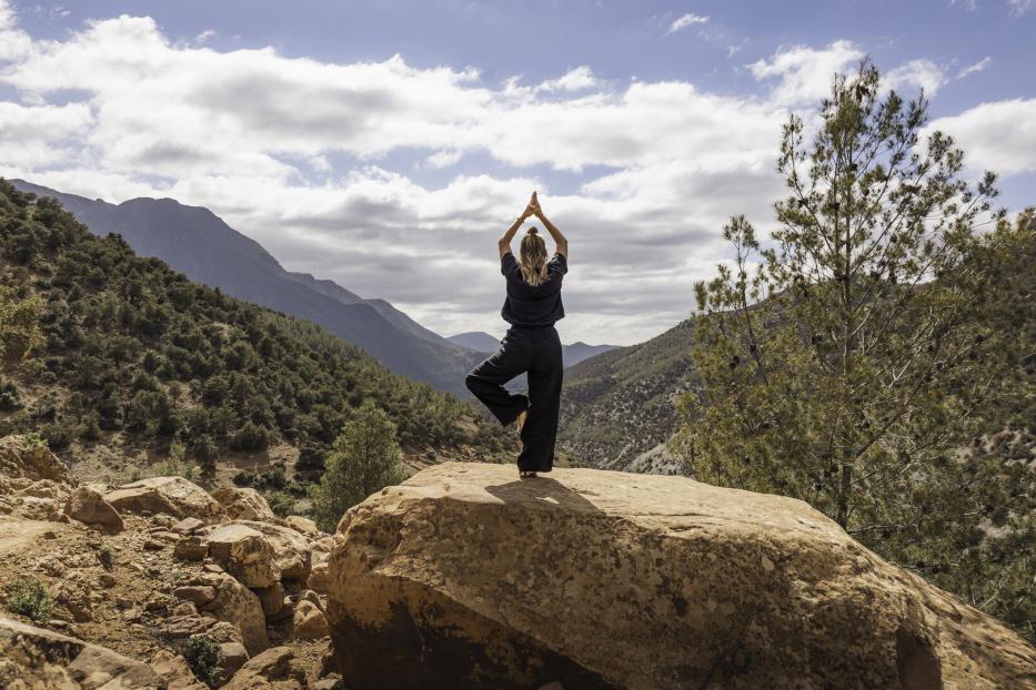 One woman practising yoga in tree pose on rock overlooking forested mountains in nature in the Atlas Mountains, Ourika Valley, Morocco