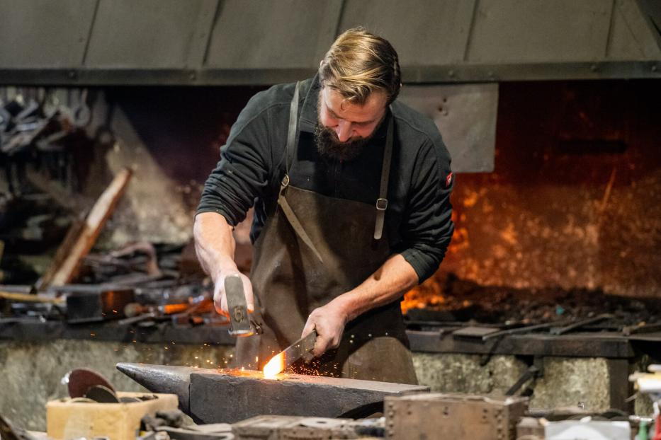 Ein Schmied mit Bart bearbeitet glühendes Metall auf einem Amboss mit Hammer.