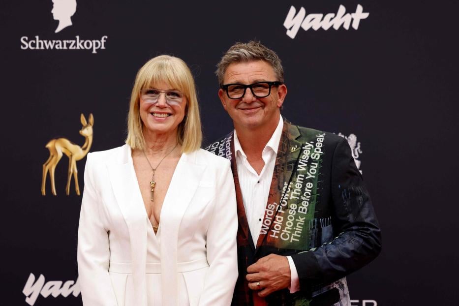 Eine Frau in weißem Anzug und ein Mann mit bedrucktem Sakko stehen lächelnd nebeneinander vor einer dunklen Wand mit Logos.