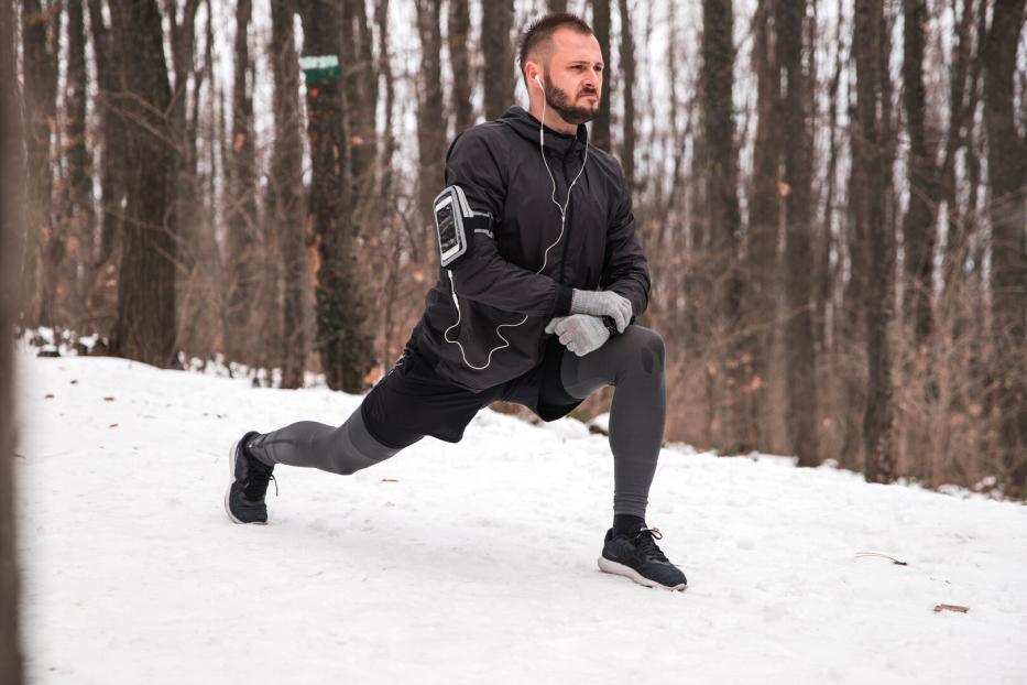 Ein Mann macht einen Ausfallschritt im verschneiten Wald und trägt Sportkleidung sowie Kopfhörer.