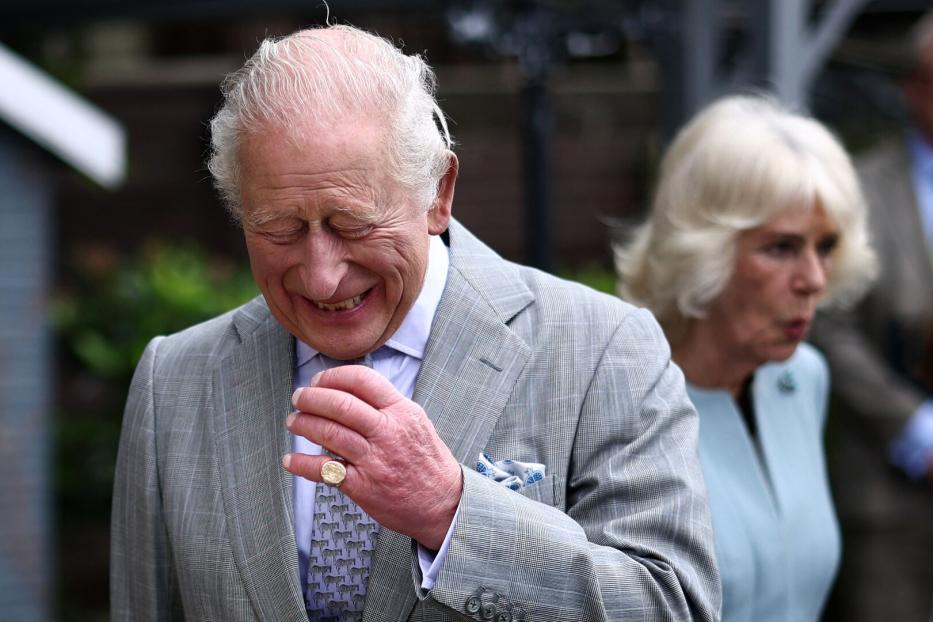 König Charles III. lächelt in einem grauen Anzug, während Camilla im Hintergrund zu sehen ist.