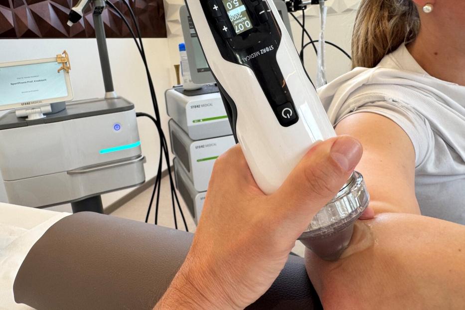 Das Bild zeigt die Anwendung der Stoßwellentherapie an einem Ellbogen, das Gerät, das die Stoßwellen erzeugt, wird direkt an den Ellbogen gehalten.