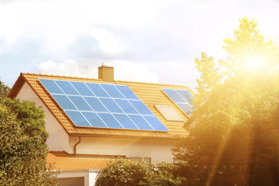 Auf einem Hausdach mit roten Ziegeln sind mehrere Solarpanels installiert, Sonnenstrahlen scheinen darauf.