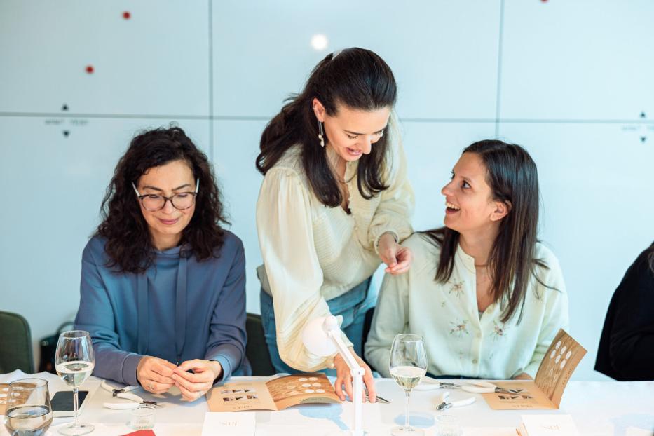 Drei Frauen sitzen an einem Tisch und arbeiten gemeinsam an Schmuckstücken, während eine lächelnd Anweisungen gibt.