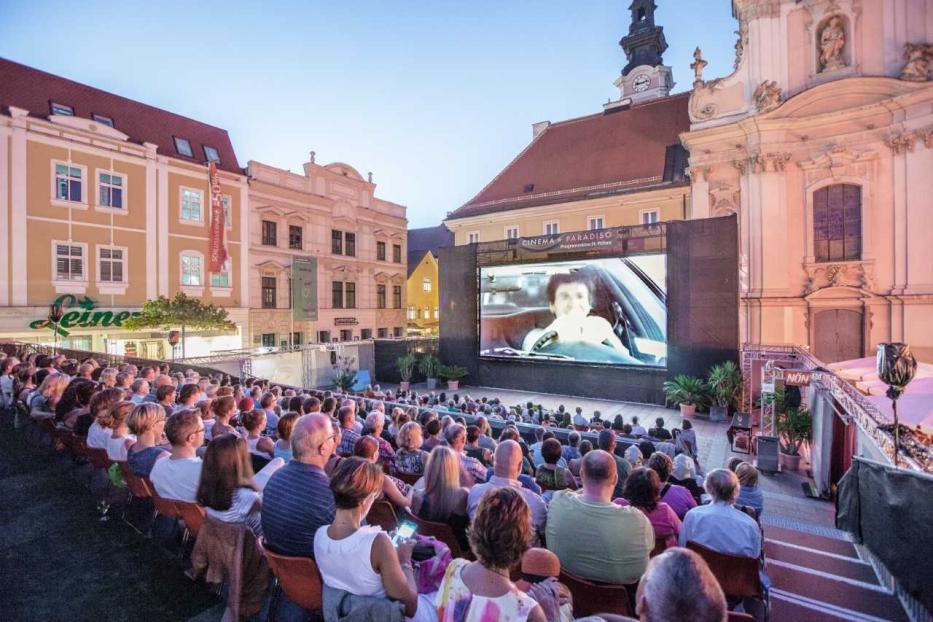 Zuschauer sitzen auf einem Platz vor einer großen Leinwand des Cinema Paradiso in St. Pölten bei einer Open-Air-Filmvorführung.
