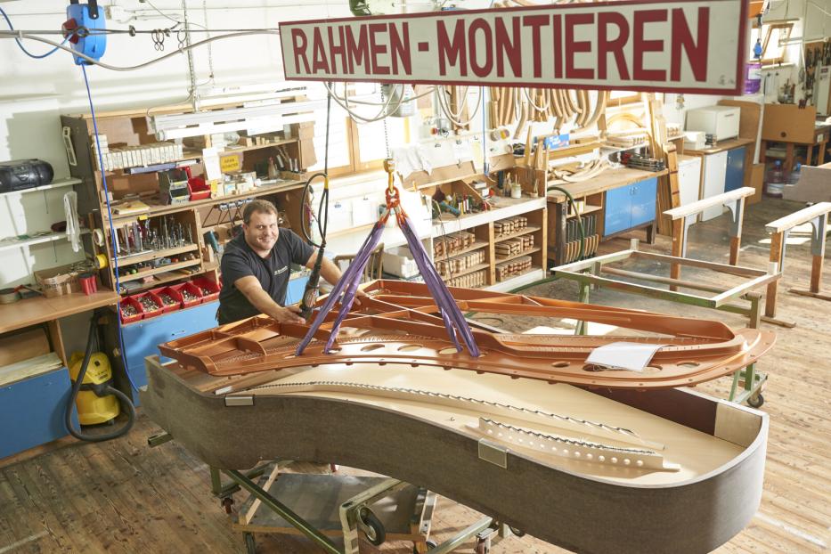 Ein Handwerker montiert in einer Werkstatt einen Flügelrahmen unter einem Schild „RAHMEN-MONTIEREN“.