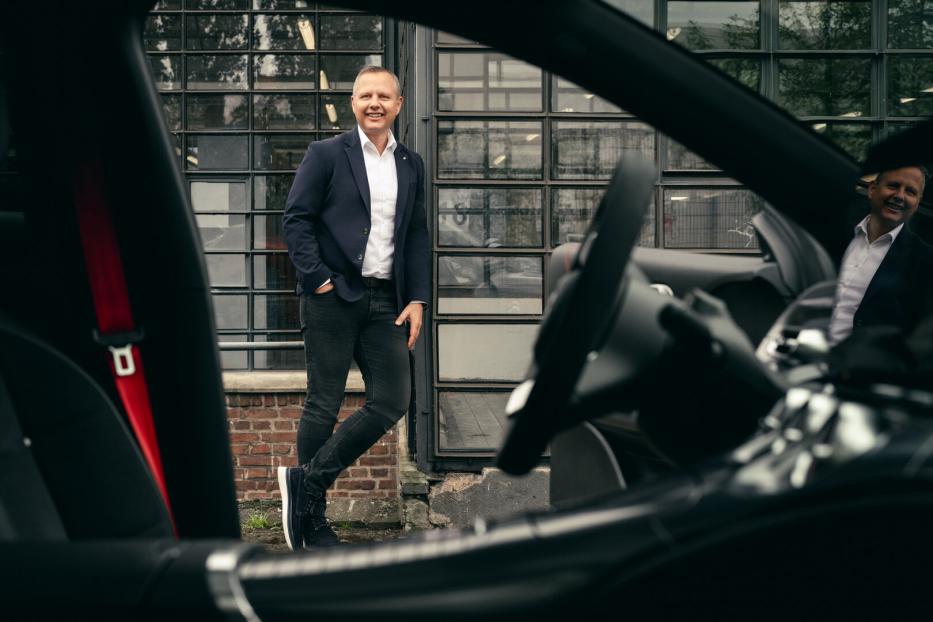 Mann im blauen Blazer steht lächelnd vor einem Auto und einem Industriegebäude.